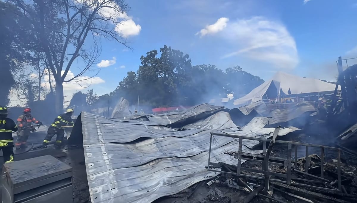 Firefighters working amid the smoking, collapsed sheet-metal remains of a large structure, with a white event tent from the attraction still standing in the background.