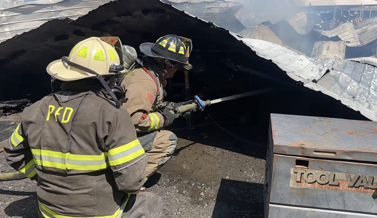 Two firefighters, one in Pleasantdale Fire Department gear, kneeling close to the debris and directing a hose stream into the blackened wreckage.