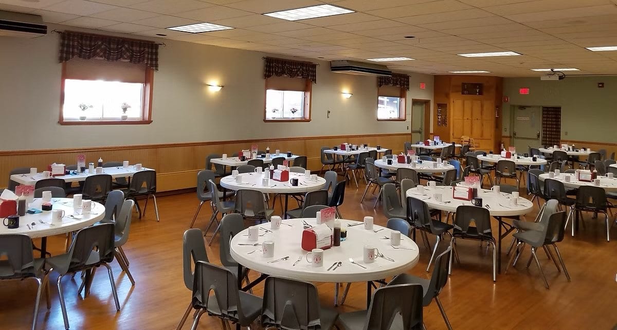 Inside the Speigletown firehouse social hall: a warm wood-floored room with round tables set with mugs and place settings, ready for a community breakfast.