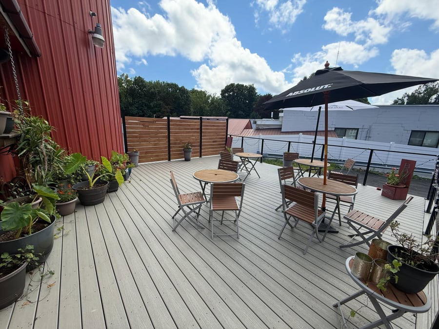The Rustic Barn Pub's newly finished deck on a sunny day: light-gray composite decking, café tables and folding chairs, a black Absolut umbrella, potted plants along the pub's red barn wall, a wood privacy screen, and a cable railing strung with string lights, with rooftops and trees beyond.