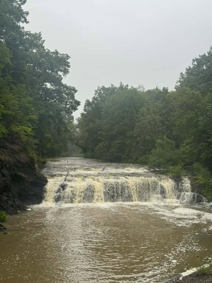 Buttermilk Falls in Schaghticoke running high after heavy rain — brown, silt-laden water spilling across the full width of the rock ledge into a swollen pool, under a grey sky.