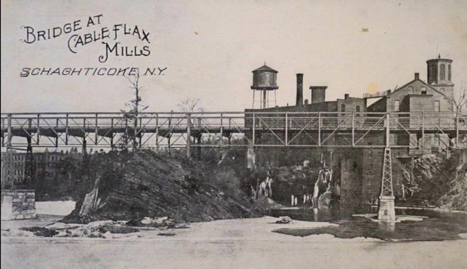 A black-and-white postcard captioned 'Bridge at Cable Flax Mills, Schaghticoke, N.Y.' An iron truss bridge spans the rocky Hoosic River gorge; brick mill buildings with a water tower and smokestacks stand on the right bank, with more mill buildings visible across the gorge at left.